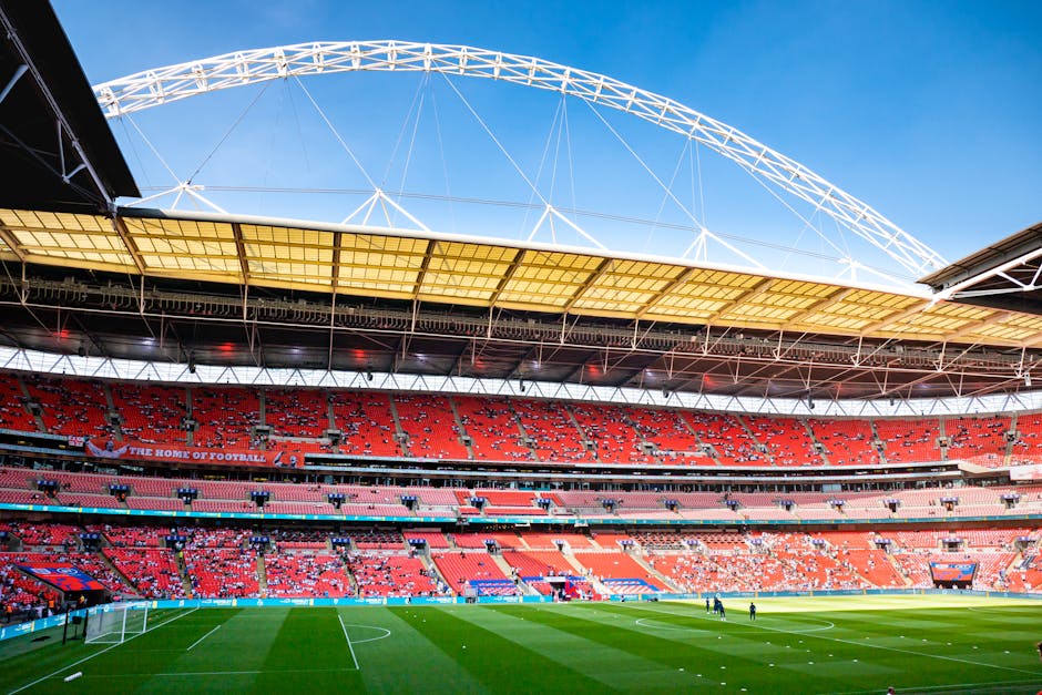 A wide view of a football stadium shows a lush green pitch with freshly cut grass, marked with white boundary and goal lines. Surrounding the pitch are multiple tiers of red stadium seats, some with s