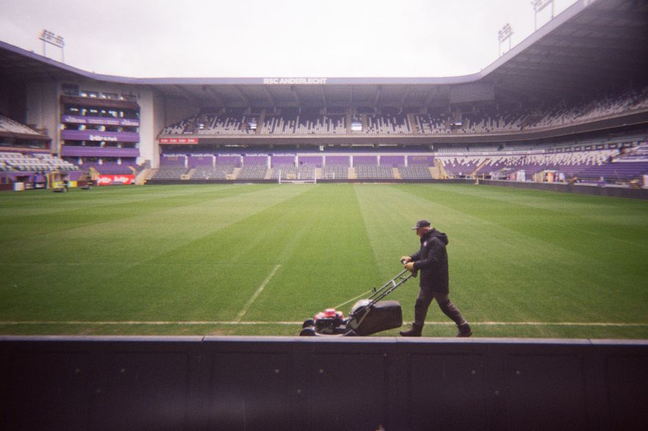 A wide view of a football stadium shows a lush green pitch with freshly cut grass, marked with white boundary and goal lines. Surrounding the pitch are multiple tiers of red stadium seats, some with s