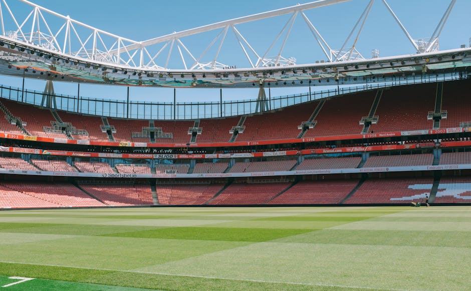 The image depicts an empty football stadium with a well-maintained grass pitch featuring alternating shades of green in a striped pattern. Surrounding the pitch, there are multiple tiers of red plasti