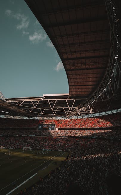 The image depicts an empty football stadium with a well-maintained grass pitch featuring alternating shades of green in a striped pattern. Surrounding the pitch, there are multiple tiers of red plasti